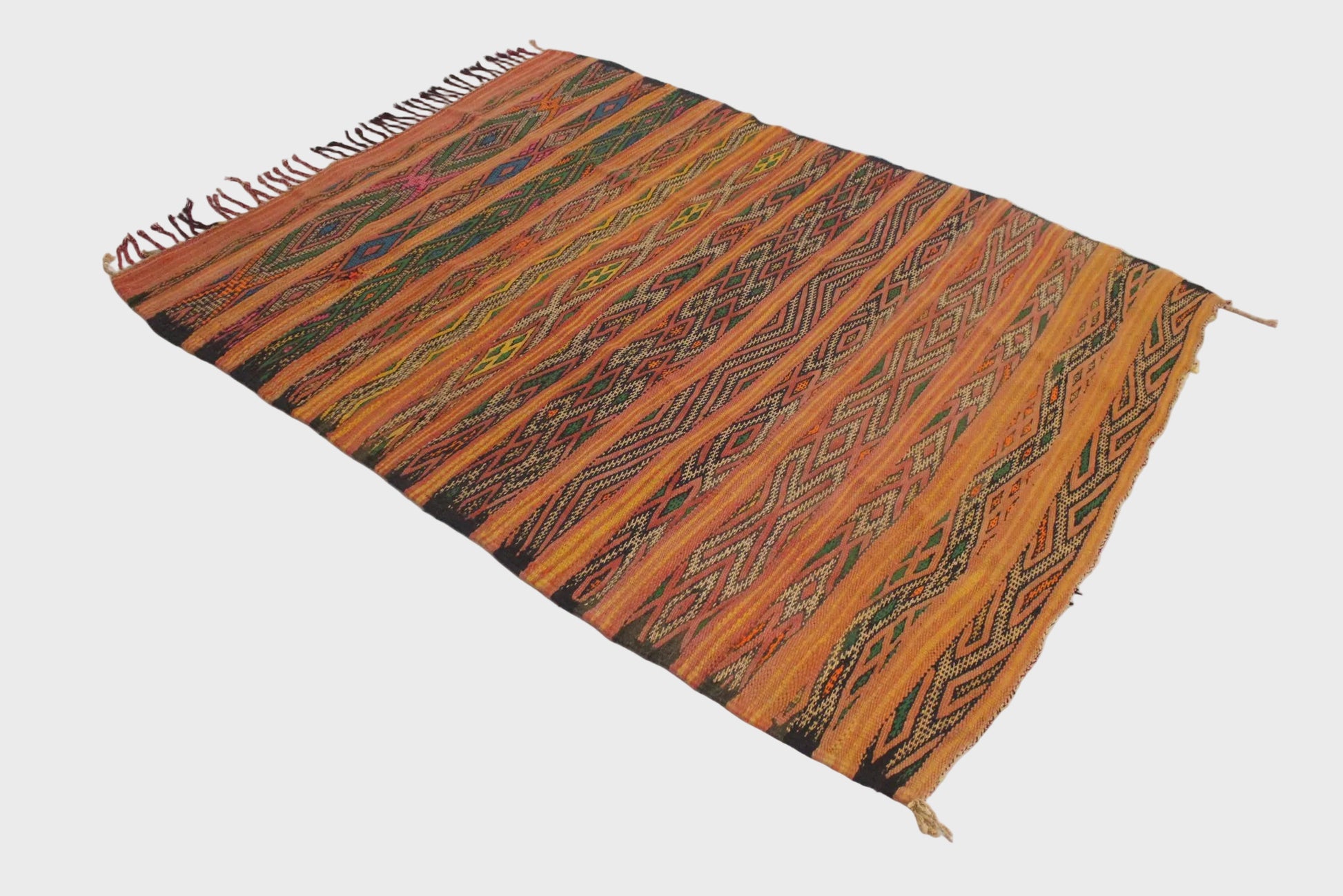 A rectangular handwoven kilim rug with a vibrant orange and brown striped pattern, featuring geometric diamond motifs and fringed edges, displayed on a plain white floor.