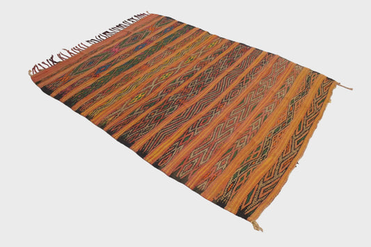 A rectangular handwoven kilim rug with a vibrant orange and brown striped pattern, featuring geometric diamond motifs and fringed edges, displayed on a plain white floor.