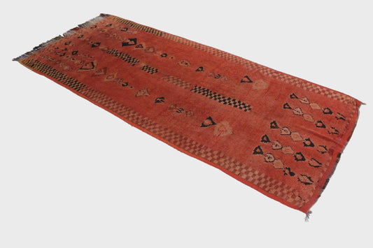 A rectangular hand-woven runner rug with a terracotta red-orange base, featuring black, beige, and brown geometric patterns and fringed edges, displayed on a light gray floor.