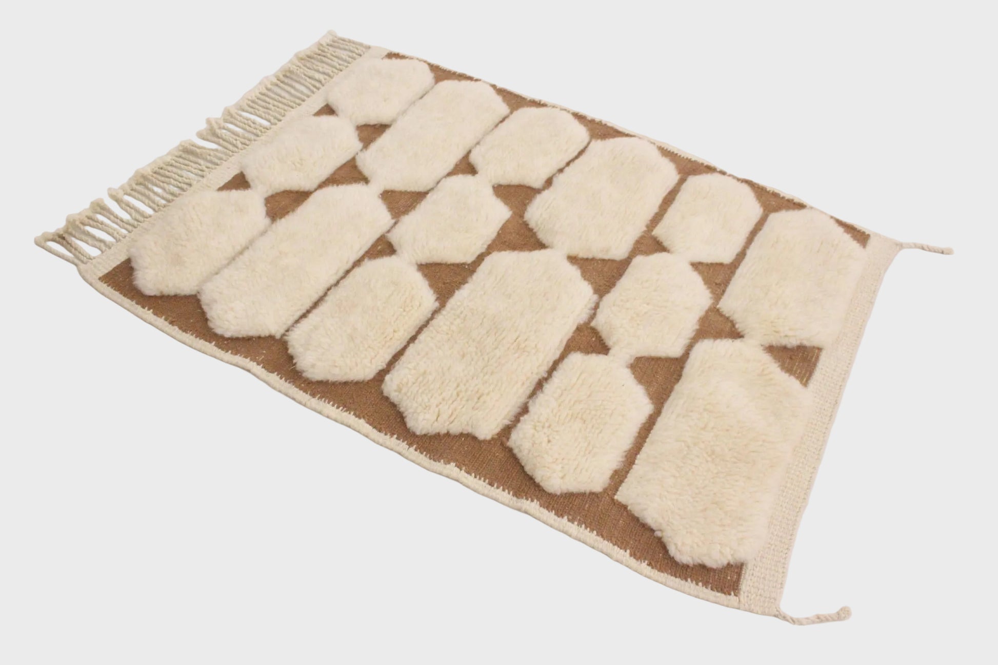 A rectangular woven rug with a geometric pattern of cream-colored raised shapes on a tan background, finished with a cream fringe on one end, shown on a plain white surface.